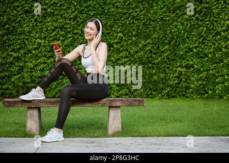 Junge Frau in Sportbekleidung, die Musik mit Kopfhörern auf dem Park hört. Sie saß auf einer Bank, ruhte sich aus und benutzte das Smartphone. Stockfoto