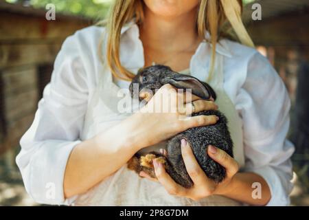 Nahaufnahme einer jungen, schönen Frau mit einem kleinen schwarzen Kaninchen. Ein hübsches, weißes Mädchen mit Strohhut und weißem Hemd lächelt und hält ein süßes Häschen in der Hand Stockfoto