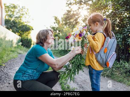 Süße Enkelin umarmt ihre glückliche Großmutter. Die Umarmungen einer älteren Frau und eines kleinen Mädchens vor dem Hintergrund von Sommerblumen, Bäumen und Sonne Stockfoto