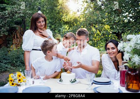 Glückliche Familie isst draußen an einem Tisch zu Mittag. Mom, Dad, zwei Söhne sind auf der offenen Veranda des Hauses oder in einem offenen Café. Sie feiern eine Geburt Stockfoto