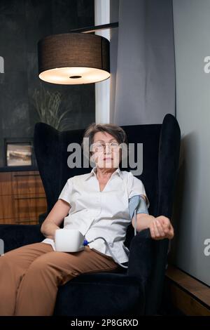 Eine ruhige alte Frau sitzt im Sessel und misst ihren Blutdruck Stockfoto