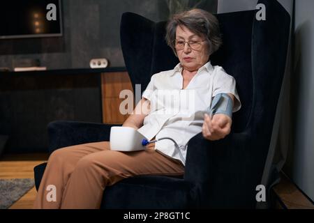 Die moderne alte Frau sitzt im Stuhl und misst ihren Blutdruck Stockfoto