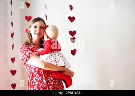 Fröhlich lächelnde Mutter und ihr Baby auf den Armen kuschelten vor Girlanden mit vielen roten Herzen. Stockfoto