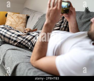 Tierbesitzer, der auf seinem Smartphone ein mobiles Spiel spielt, während sein Schoßhund zwischen den Beinen liegt und sich entspannt. Stockfoto