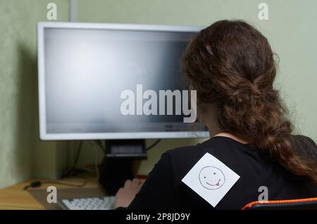 Eine Frau mit einem Aprilscherz-Zettel auf dem Rücken arbeitet an einem Computer Stockfoto