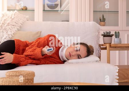Verärgerte junge Frau, die ein orthopädisches Halsband trägt und den Sender wechselt, während sie zu Hause im Wohnzimmer fernsieht Stockfoto
