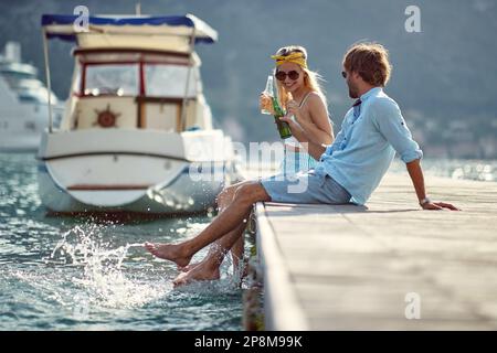 Ein erwachsenes weißes Paar sitzt auf einem Dock, redet, lächelt, lacht, spritzt Wasser mit Beinen, hat Spaß am Meer. Speicherplatz kopieren Stockfoto