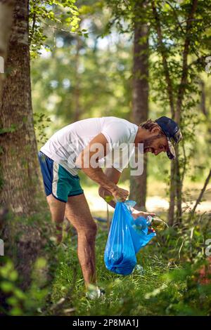 Junger Mann in weißem Hemd in einem grünen Quellwald bei sonnigem Wetter, mit blauer Plastiktüte, die Müll sammelt. Umweltschutz, Umweltschutz, Lifestyle Co Stockfoto