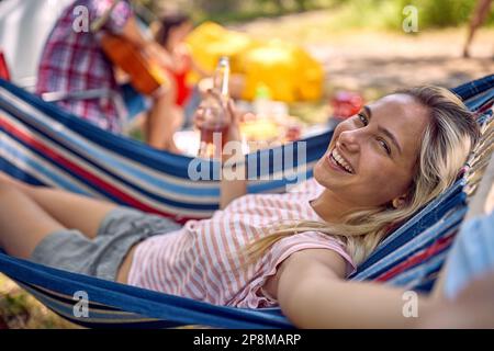 Junge, fröhliche Hipsterin, die Selfie macht, während sie in der Hängematte liegt und Bier hält. Urlaub, Zusammensein, Spaß, Lifestyle-Konzept. Stockfoto