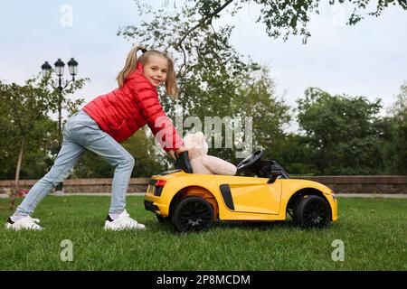 Ein süßes kleines Mädchen, das mit einem Spielzeugbären spielt, und ein Kinderauto auf dem Parkplatz Stockfoto