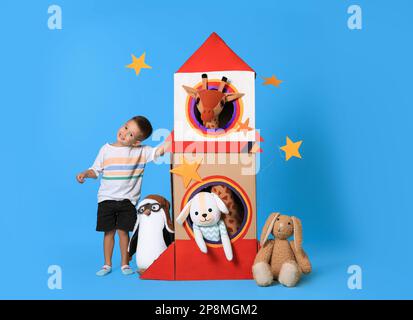 Süßer kleiner Junge, der mit einer Papprakete und weichen Spielzeugen auf hellblauem Hintergrund spielt Stockfoto