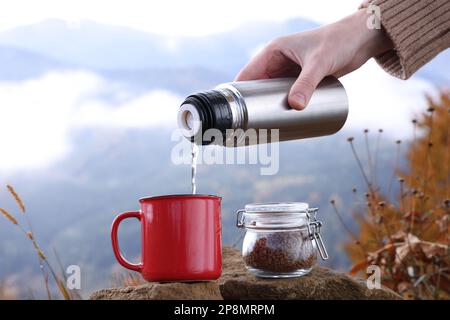 Frau, die heißes Wasser aus der Thermoskanne in eine Tasse mit Instantkaffee in den Bergen gießt, schließt Stockfoto