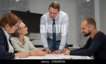 Ein weißer Mann steht und ändert eine Zeichnung, drei Kollegen sitzen an einem Tisch und hören ihm zu. Stockfoto