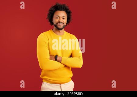 Ein bärtiger, gutaussehender Mann, isoliert auf rotem Hintergrund, mit gekreuzten Armen, schaut in die Kamera, männlicher Unternehmer in Freizeitkleidung Stockfoto