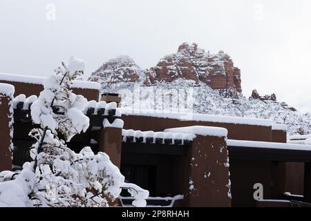 Ein Gebäude im Pueblo-Stil im Vordergrund und rote Felsenberge im Hintergrund, alle mit Schnee bedeckt nach einem Wintersturm in Sedona, Arizona. Stockfoto