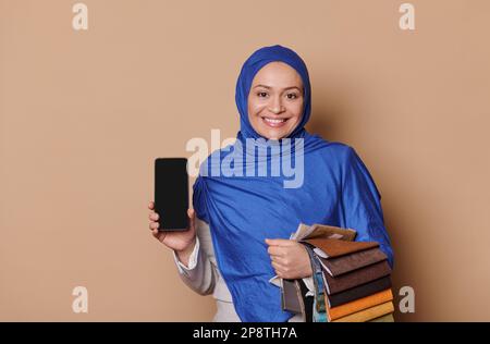 Eine hübsche muslimische Frau in blauem Hijab, Vertriebsmanager im Möbelgeschäft, zeigt ein Smartphone mit leerem Bildschirm für die mobile App Stockfoto