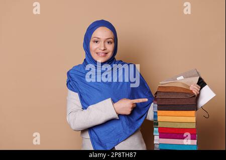 Selbstbewusste muslimische Frau in blauem Hijab, präsentiert Katalog von Polsterstoffen, lächelt in die Kamera, beiger Hintergrund Stockfoto