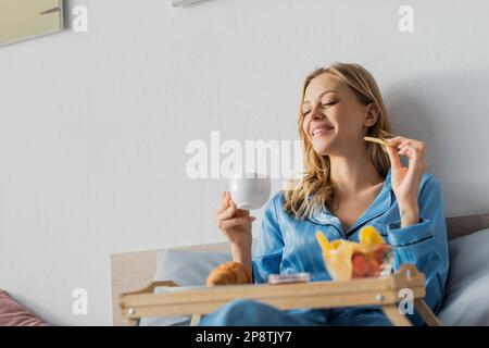 Eine freudige Frau im Pyjama hielt eine Tasse Kaffee und getrocknete Mango neben dem Tablett, während sie im Bett frühstückte Stockfoto