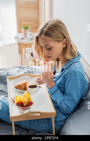 Blonde junge Frau im Pyjama hält eine Tasse Kaffee und frische Erdbeeren neben dem Tablett, während sie im Bett frühstückt, Bild von Stock Stockfoto