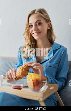Hübsche Frau in blauem Pyjama, die frische Croissants neben dem Tablett hält, während sie im Bett frühstückt, Stockbild Stockfoto