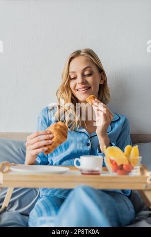 Glückliche Frau im Pyjama, die frisches Croissant neben dem Tablett isst, während sie im Bett frühstückt, Bild von Stock Stockfoto