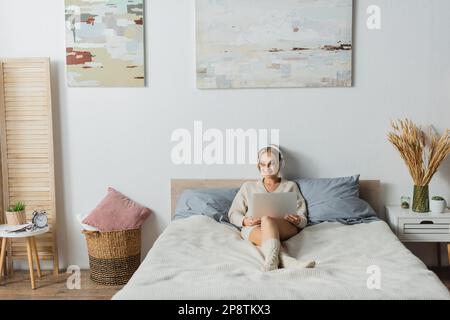 Volle Länge einer fröhlichen jungen Frau in kabellosen Kopfhörern mit Laptop im modernen Schlafzimmer, Stockbild Stockfoto