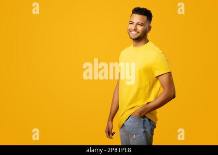 Ein hübscher, lächelnder indischer junger Mann in einem gelben, lässigen T-Shirt, der auf gelbem Hintergrund posiert, charismatischer Typ mit einem zahnhaften Lächeln, der zur Seite steht Stockfoto