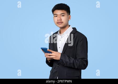 Junger, gutaussehender asiatischer Geschäftsmann, der mit einem Smartphone in der Hand lächelt, isoliert auf blauem Hintergrund Stockfoto