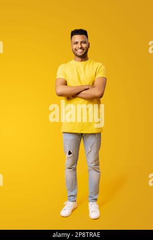Großes Porträt eines selbstbewussten, glücklichen jungen Indianers 20s in einem gelben T-Shirt, der Hände gekreuzt hat, isoliert auf einfarbigem gelben Hintergrund, ausländischer männlicher Student Stockfoto