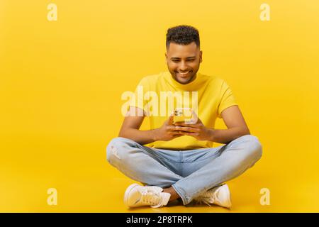 Süchtig nach Nachrichten lächelnder junger indischer 20s-Mann mit Smartphone in den Händen, der in Lotus-Pose isoliert auf Gelb sitzt. Ein gut aussehender optimistischer Kerl, der eine mobile App benutzt, online chattet, ein Spiel spielt Stockfoto