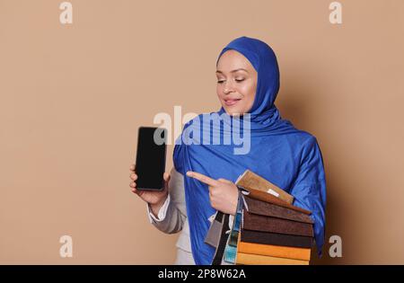 Angenehme muslimische Vertriebsleiterin im Hijab, hält Stoffproben und zeigt Smartphone mit leerem Bildschirm für mobile Apps Stockfoto