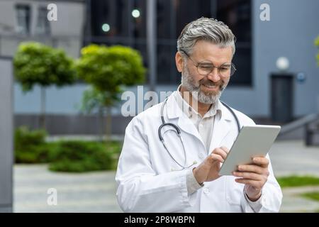 Porträt eines lächelnden, erfolgreichen, grauhaarigen, männlichen Hausarztes in einem weißen Kittel und mit einem Stethoskop, der vor der Klinik steht, ein Tablet hält, online arbeitet, chattet, online telefoniert. Stockfoto