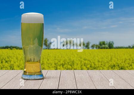 Ein Glas helles Bier auf dem Tisch mit Blick auf das Feld mit gelben Blumen und blauem Himmel. Stockfoto