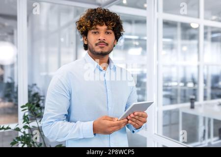 Porträt eines jungen hispanischen Studenten in einem blauen Hemd, der im Büro des Campus mit einem Tablet in der Hand stand und ernsthaft in die Kamera schaute. Stockfoto