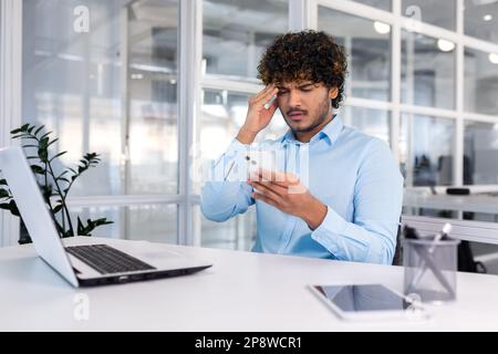 Probleme mit dem Konto, der Anwendung, dem Online-Shopping, einer fehlerhaften Nachricht. Ein junger lateinamerikanischer Mann sitzt an einem Schreibtisch im Büro, hält seinen Kopf fürchterlich fest und schaut sich traurigerweise sein Telefon an. Stockfoto