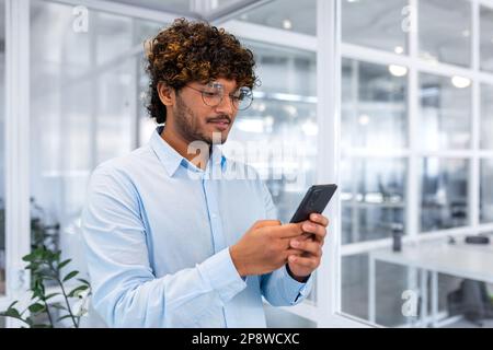 Ein junger Inder steht im Büro und telefoniert, wählt Nachrichten, schreibt SMS, bezahlt online, bestellt, Einkaufen. Stockfoto