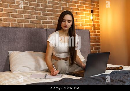 Glückliche schöne Frau sitzt auf dem Bett, arbeitet an einem Laptop, macht Notizen in einem Notizbuch. Stockfoto