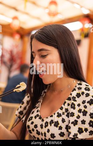 Weibliches Model im Restaurant isst mit Stäbchen, Lebensstil einer jungen Frau mit schwarzem, glattem Haar, traditionelles asiatisches Essen und Küchenutensilien Stockfoto