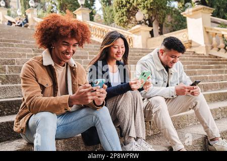Junge Freunde aus einer Gruppe verschiedener Rassen, die ihre Mobiltelefon-App im Freien nutzen und lächeln. Drei glückliche, multiethnische Teenager, die mit ihrem Handy Spaß beim Spielen haben. Kommunikationskonzept. Hochwertiges Foto Stockfoto