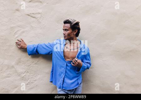 Junger, stylischer Kerl in einem blauen Hemd, der draußen an der Wand steht Stockfoto