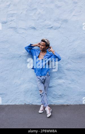 Ein Typ in einem blauen Hemd, der die Wand nach draußen neigt. Die ganze Länge eines jungen, gutaussehenden Mannes, der sich an einer blauen Wand entspannt. Stockfoto