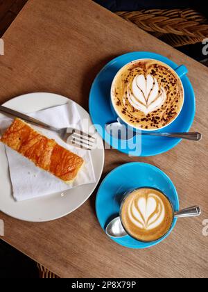 Blick von oben auf dekorierte Tassen Kaffee (Kaffeekunst) und ein Stück Zitronenkuchen Stockfoto