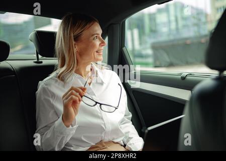 Lächelnde Geschäftsfrau Managerin, die an einem Laptop arbeitet, auf dem Rücksitz sitzt und auf das Fenster schaut Stockfoto