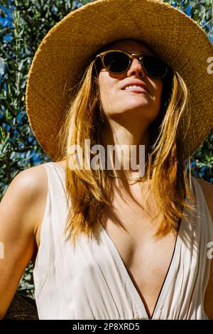Eine Frau mit Sonnenbrille und Sonnenhut steht in einem Sonnenkleid vor üppigem Laub. Stockfoto
