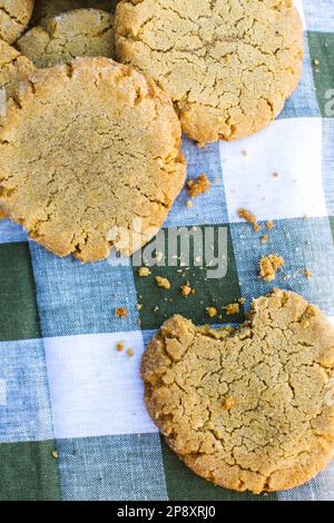 Nahaufnahme eines Haufens frisch gebackener Erdnussbutter-Kekse mit einem Bissen aus einem und Krümeln auf der Gingham-Tischdecke. Stockfoto