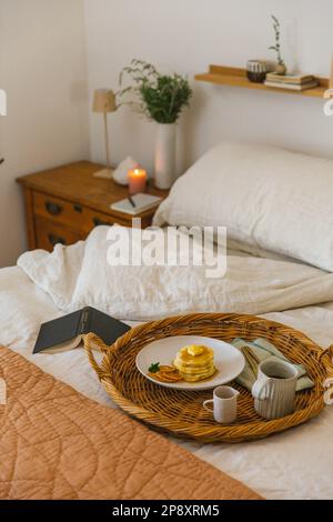 Ein perfektes Frühstück im Bett wird auf einem gewebten Tablett auf weicher Bettwäsche mit Kerze im Hintergrund serviert. Stockfoto