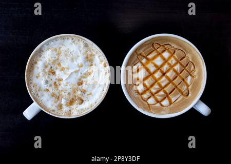 Zwei weiße Tassen nebeneinander mit Latte-Getränken in weißen Tassen auf einem dunklen Tisch, Draufsicht Stockfoto