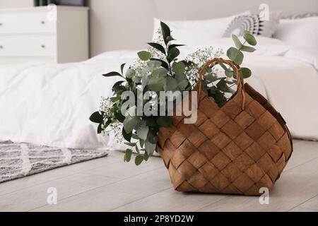 Stilvoller Korb aus Korb mit frischen Eukalyptuszweigen und Blumen auf dem Boden im Schlafzimmer Stockfoto