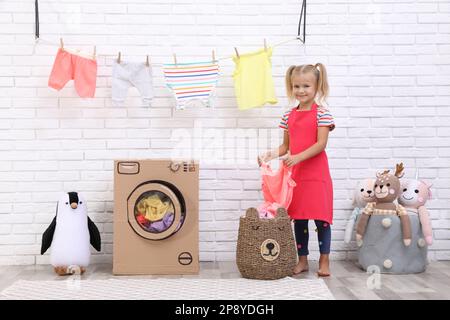 Ein kleines Mädchen, das saubere Wäsche in der Nähe der Spielzeugpappe-Waschmaschine im Haus hält Stockfoto