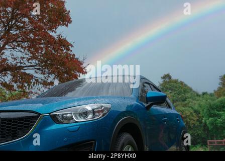 Blauer SUV mit Regentropfen vor grünem Wald und Regenbogenhimmel. Parkplatz im Freien. Roadtrip. Abenteuerreisen Stockfoto